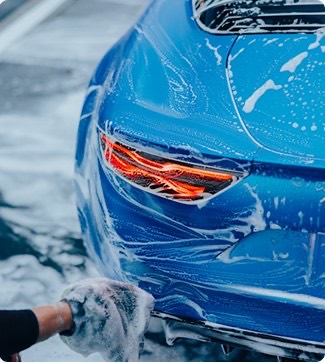 Close-up of a blue car's headlight being hand-washed with soap suds.
